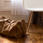 A khaki canvas duffel bag on a light wooden floor, next to a white plastic chair with metal legs.