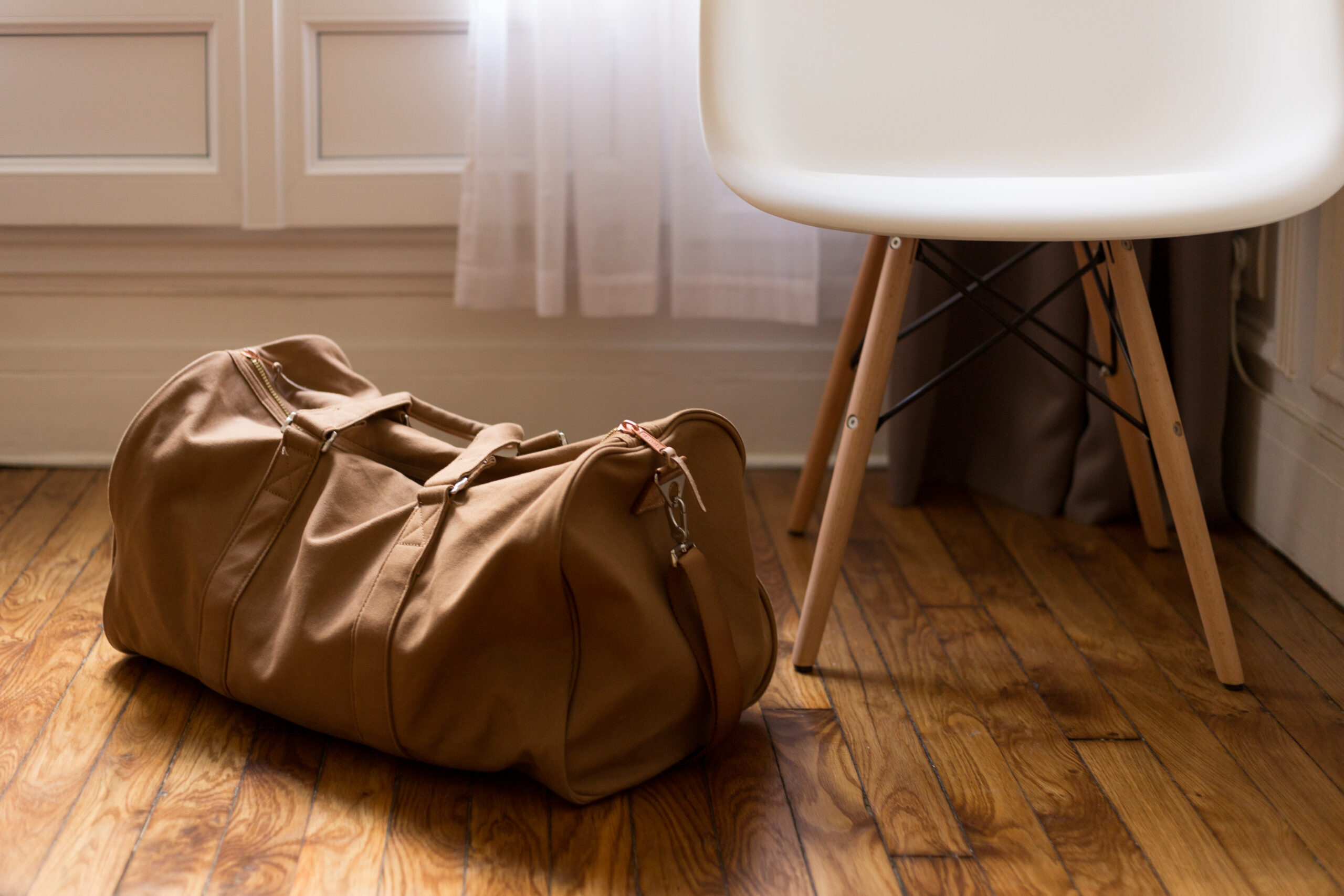 A khaki canvas duffel bag on a light wooden floor, next to a white plastic chair with metal legs.