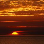 A sun hanging low on the horizon, creating an deep orange sunset behind clouds.